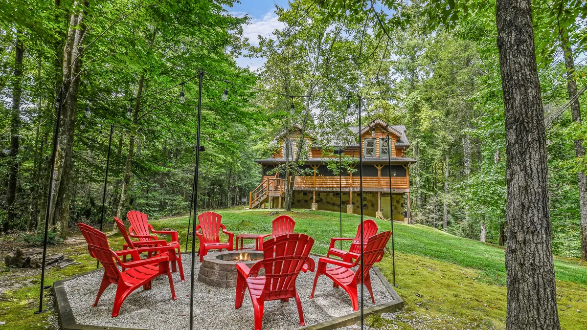 A cozy fire pit surrounded by red Adirondack chairs and framed by a rustic wooden house and tall trees. The scene is inviting, ideal for shared stories, marshmallow roasting, and stargazing under the open sky.