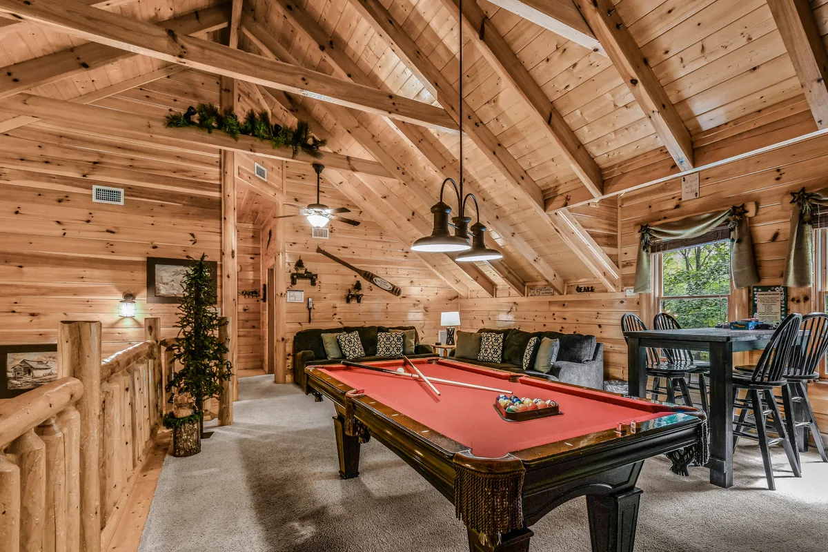 A stylish game loft featuring a pool table in the center, a comfortable couch with decorative pillows, exposed wooden beams, and a window offering a scenic woodland view. The setting is open and inviting, ideal for both active play and relaxed socializing.