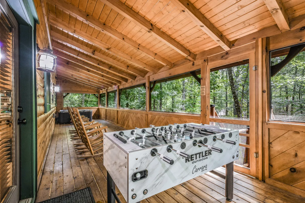 An exterior porch scene with a foosball table and wooden rocking chairs set among sturdy wooden beams, offering an expansive outdoor view of the surrounding trees. The image radiates a sense of friendly relaxation and fun, perfect for gatherings.