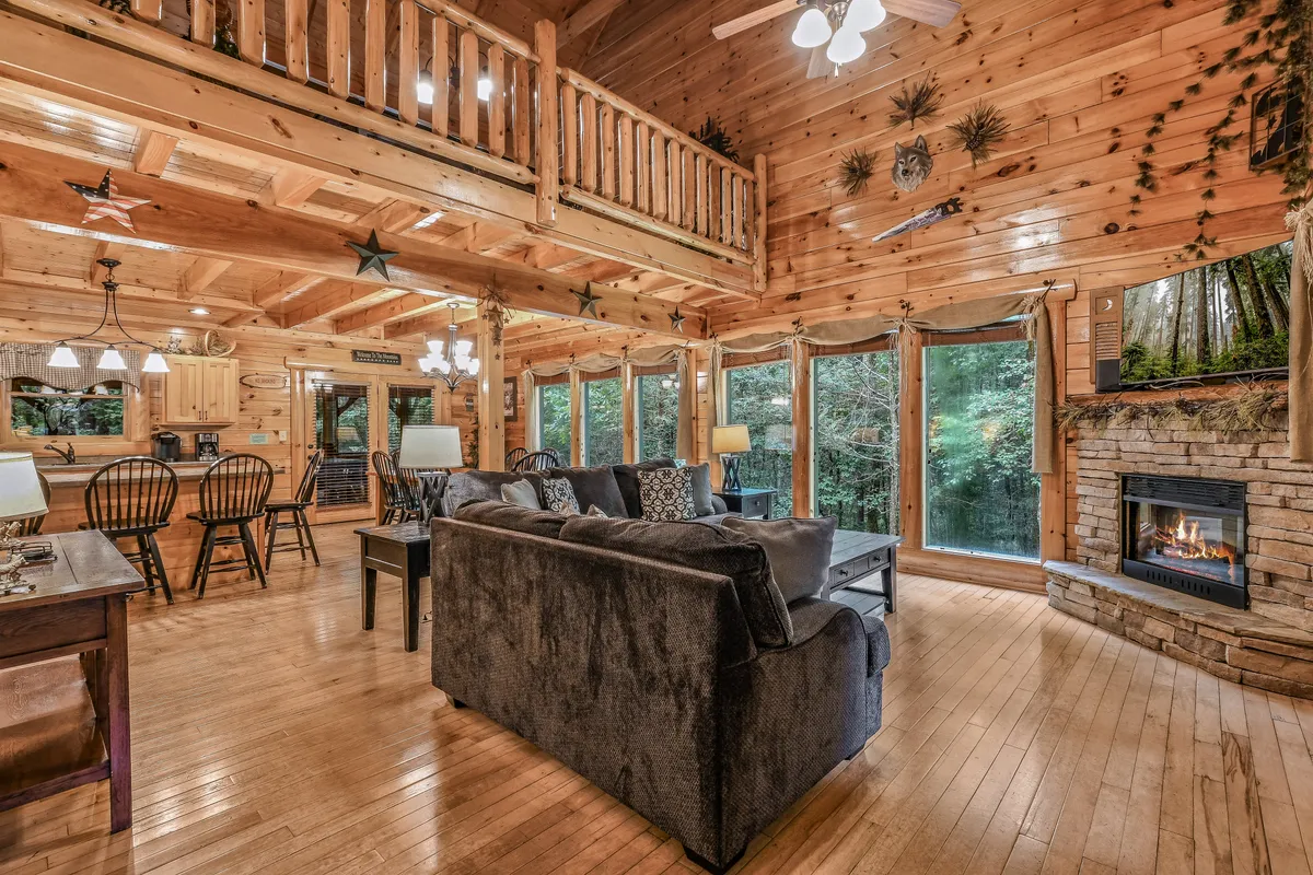 This interior image presents a warm and inviting living space featuring a prominent stone fireplace, large windows allowing natural light and views of the outdoors, rustic wooden beams overhead, plush sofas, and a dining area in the background. The arrangement highlights spaces for gathering, relaxation, and shared meals in an open-plan layout.