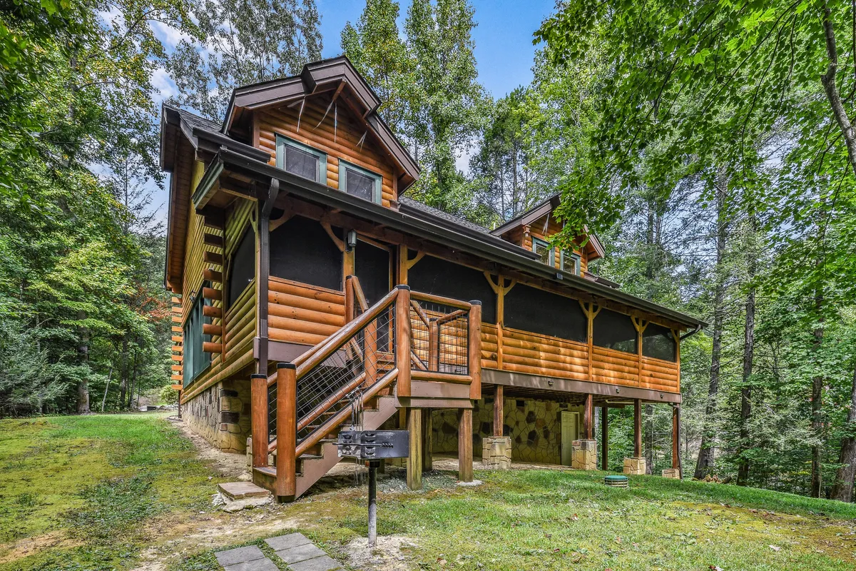 A rustic log cabin exterior nestled amidst vibrant green foliage, with inviting wooden stairs leading to a welcoming porch. The natural setting highlights the cabin’s seamless integration with its lush surroundings.