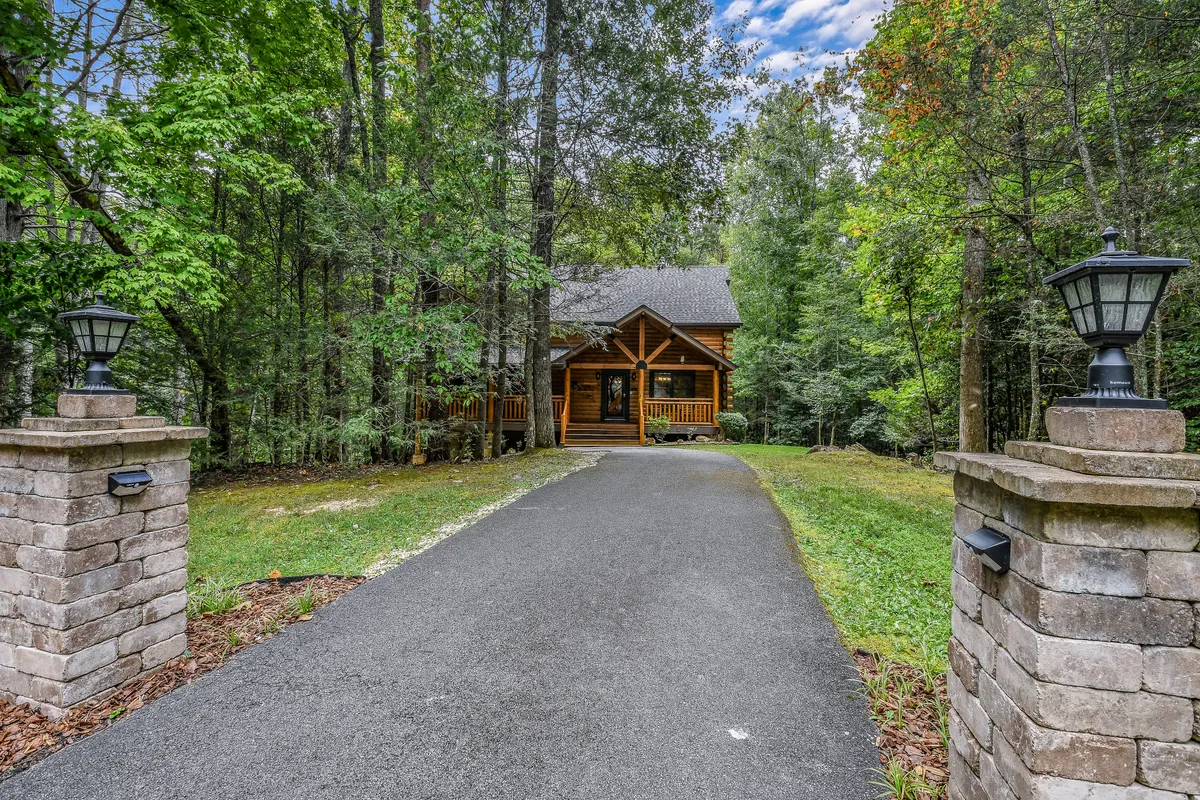 A charming log cabin with a spacious driveway and lush greenery all around. The wide front porch hints at a relaxed, welcoming atmosphere, inviting guests to gather outside for morning coffee or evening chats surrounded by nature.