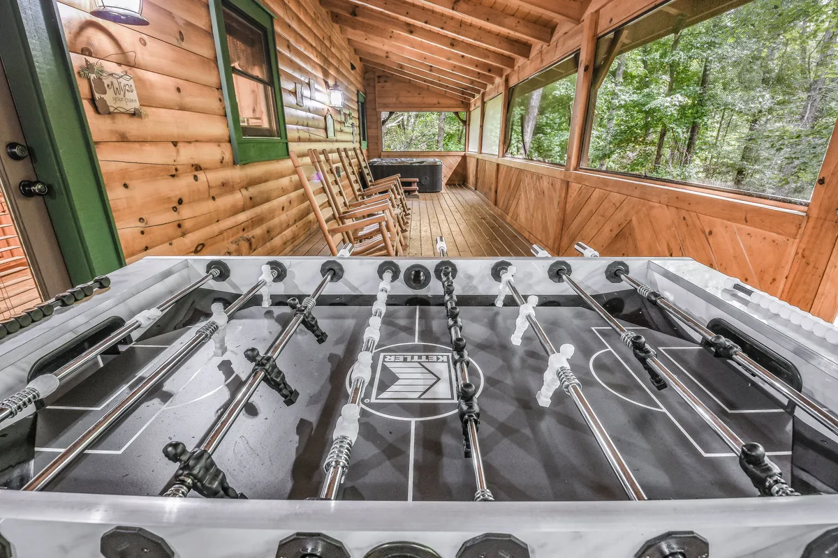 This interior scene features a foosball table in a room with large windows overlooking the trees, wooden rocking chairs, and a casual, friendly atmosphere. It suggests playful family competition mixed with opportunities for relaxation and enjoying woodland views.