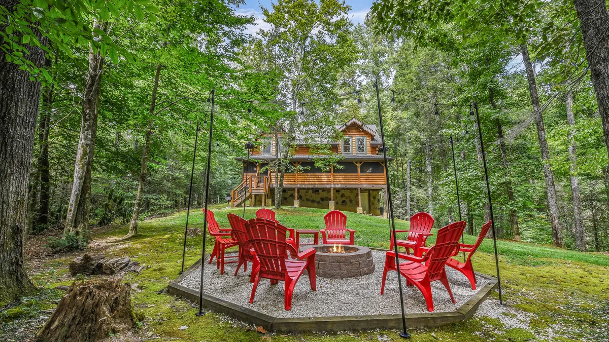 This image captures a cozy fire pit surrounded by red Adirondack chairs, set against a backdrop of a wooden cabin and towering trees. The ambiance is communal and inviting, with the scene clearly designed for outdoor gatherings, storytelling under the stars, and classic campfire moments like roasting marshmallows.