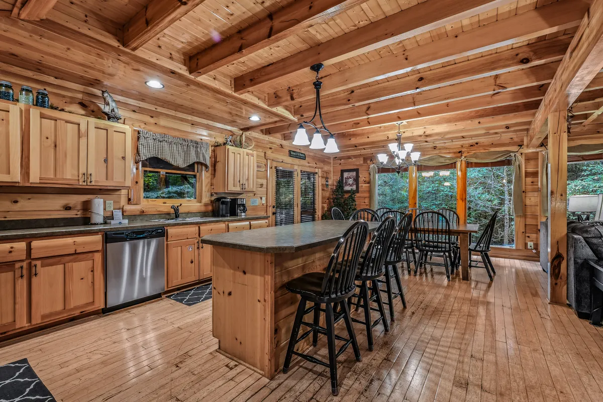 A bright, open interior with a large kitchen island featuring seating, wooden cabinetry, a dining area with a table set for gatherings, and expansive windows that bring the outdoors in. The image conveys a communal atmosphere ideal for cooking, dining, and connecting.