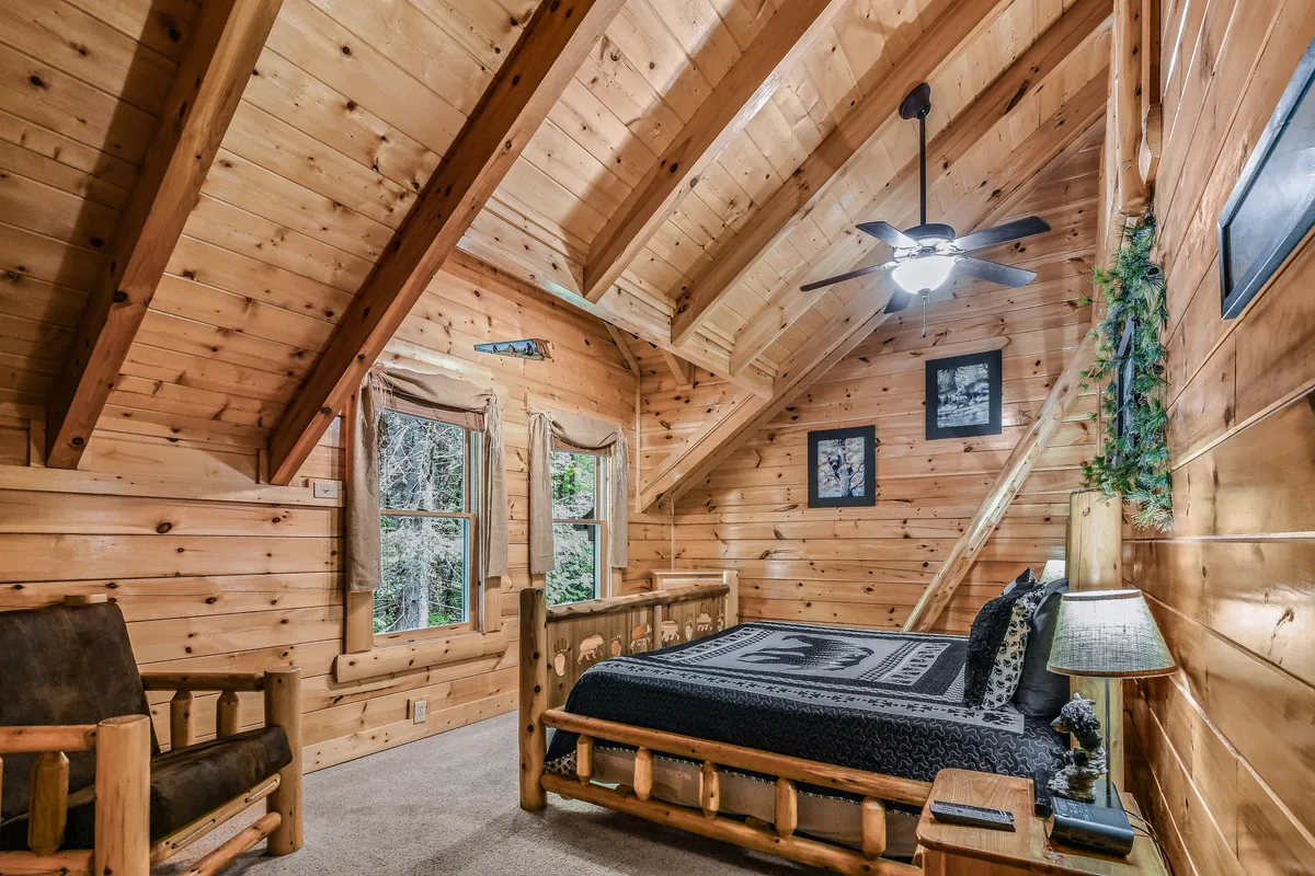 A warm, rustic bedroom with a wooden bed, large windows for abundant natural light, a ceiling fan, and wood accents throughout. The room feels cozy and inviting, with gentle sunlight illuminating the peaceful setting.