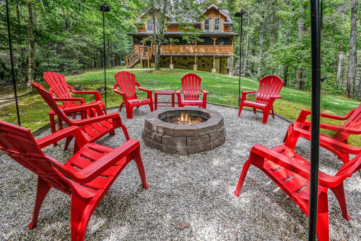 A cozy outdoor fire pit surrounded by bright red chairs sits nestled in a wooded setting, with the inviting log cabin visible in the background. The scene suggests a welcoming space for groups to gather and share stories under the stars, with nature providing a serene backdrop to the experience.