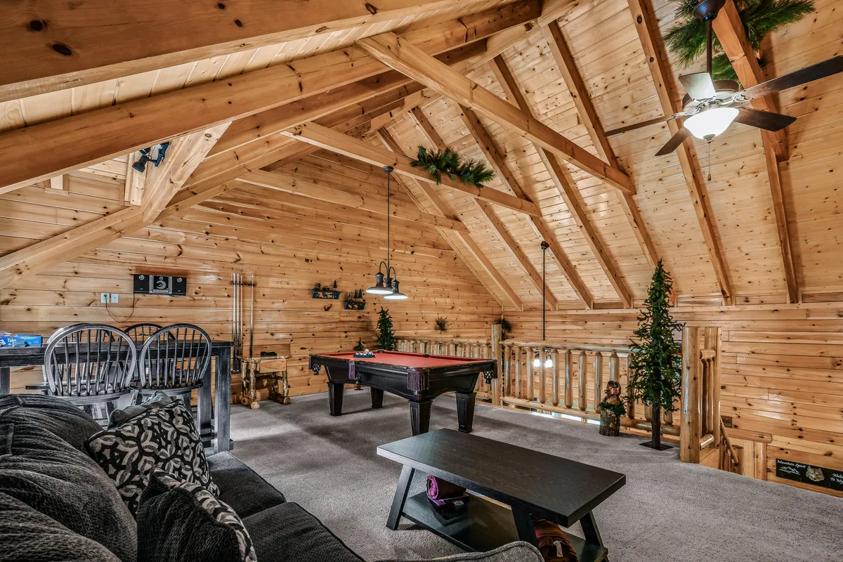 A lively play zone featuring a pool table as the focal point, surrounded by a cozy seating area, rustic wooden beams, and decorative plants. The space is arranged for group activities, movie nights, or simply relaxing between games.