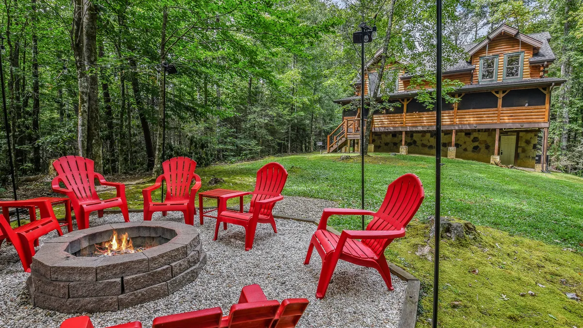 A cozy outdoor fire pit area with red chairs arranged around the fire, set beside the wooden cabin. The scene invites guests to gather, share stories, and enjoy evenings under the stars in a communal, friendly atmosphere.