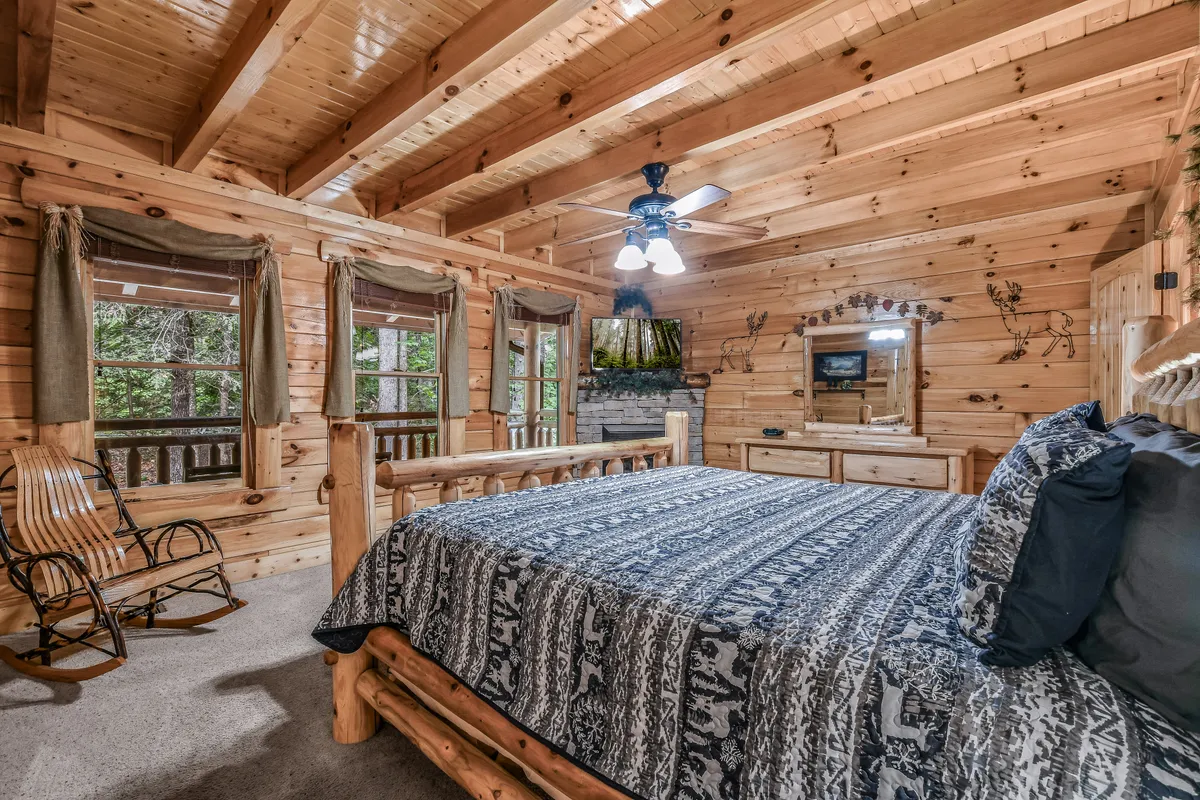 A tranquil king bedroom with a log bed, wooden rocking chair, large windows showcasing woodland views, a decorative mirror, and a television. The ambiance is inviting, with natural light filtering through the windows and warm wooden accents throughout.