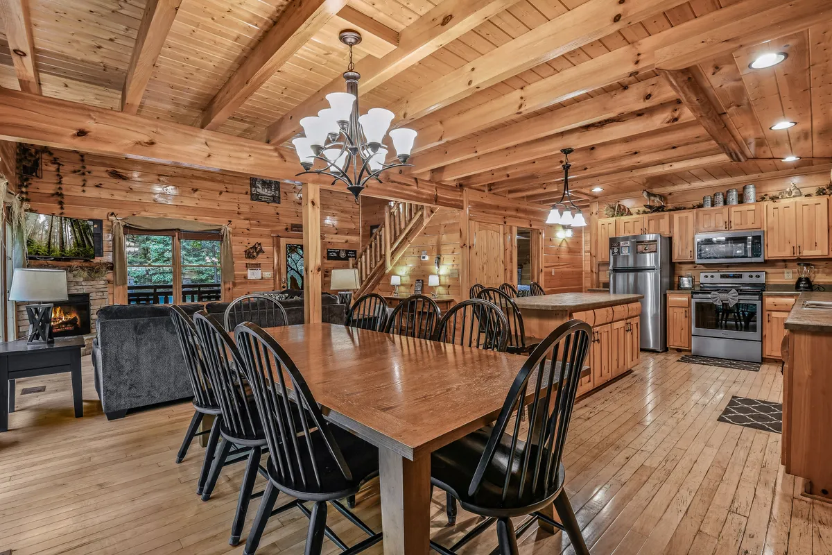 A wide view of the home’s heart: a substantial wooden dining table under rustic beams, a modern kitchen with appliances ready for meal prep, a cozy stone fireplace, and a staircase leading up. The arrangement foregrounds the dining table as a centerpiece for gathering, complemented by the warmth of the fireplace and the accessibility of the kitchen.