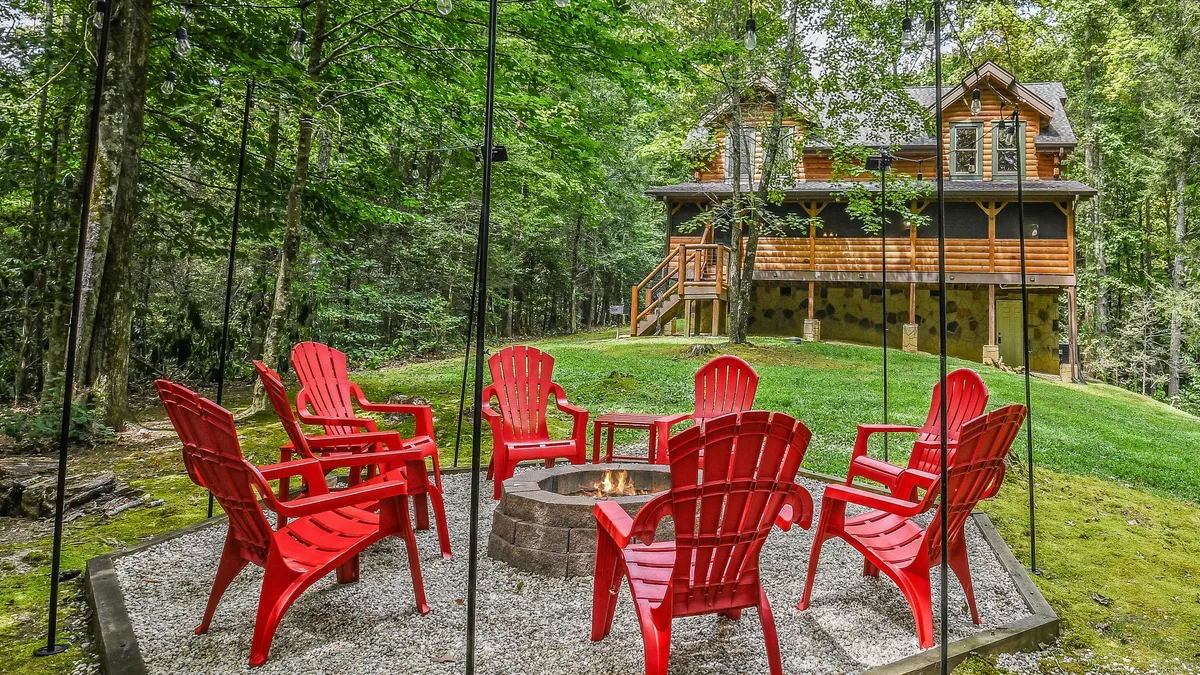 The image showcases the exterior of a rustic wooden cabin nestled among lush Smoky Mountain trees. In the foreground, a circle of vibrant red chairs surrounds a glowing fire pit, setting the scene for cozy gatherings. The soft light of sunset bathes the setting in a warm, inviting glow, highlighting the tranquil mountain ambiance and hinting at evenings spent sharing stories, roasting s'mores, and stargazing.