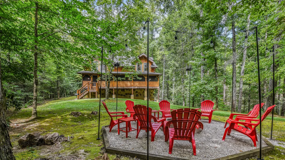 A welcoming outdoor scene featuring a cluster of red Adirondack chairs set beside a wooden cabin, surrounded by lush greenery. The setup invites guests to gather around a fire, enjoy casual conversations, and take in the serenity of the wooded landscape.